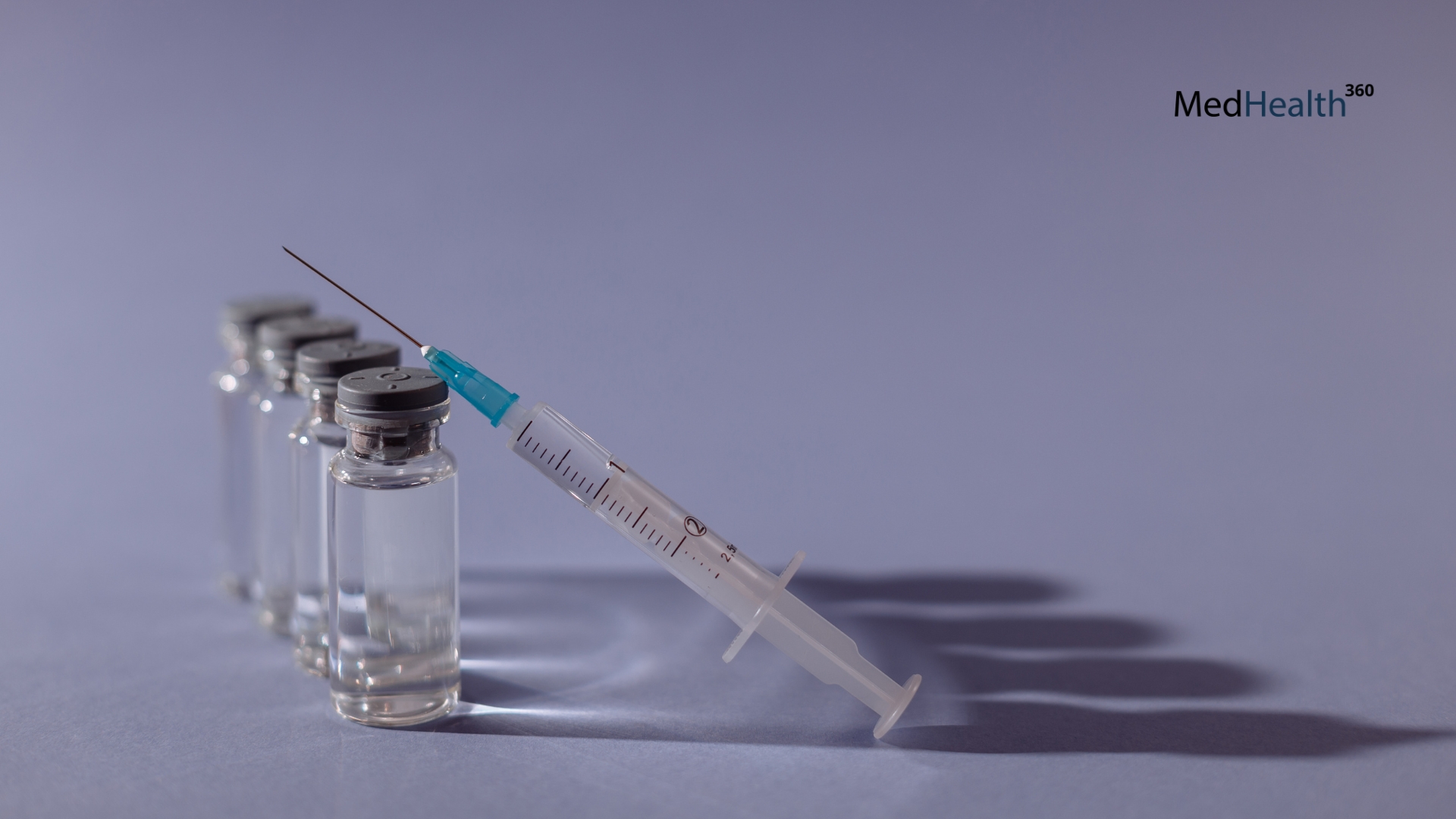 Several vials with silver caps stand in a row behind a syringe with a needle